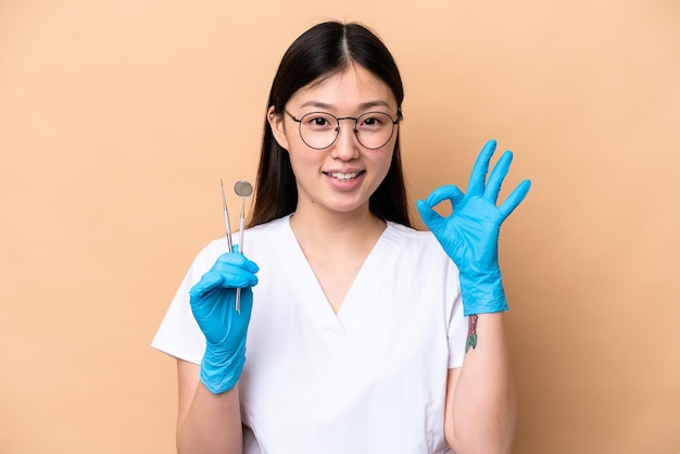 Dentist Chinese woman holding tools isolated on beige background showing ok sign with fingers