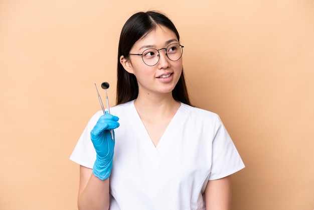 Dentist Chinese woman holding tools isolated on beige background thinking an idea while looking up