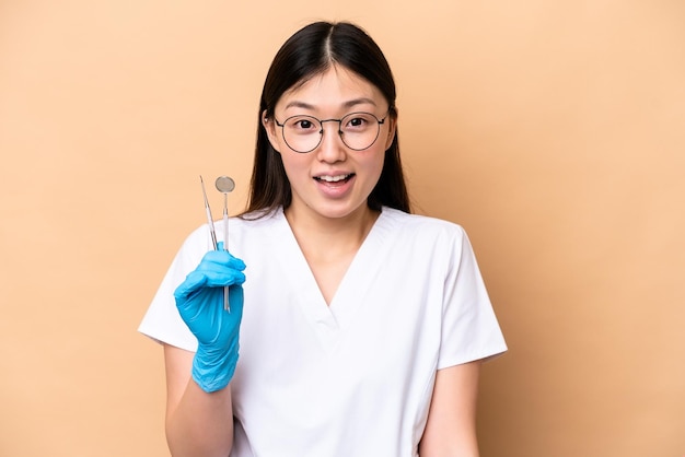 Dentist Chinese woman holding tools isolated on beige background with surprise facial expression