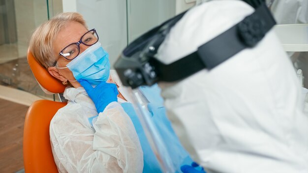 Dentist doctor in coverall preparing patient for dental surgery lighting the lamp in stomatological office during global pandemic. Medical team wearing protection suit, face shield, mask and gloves