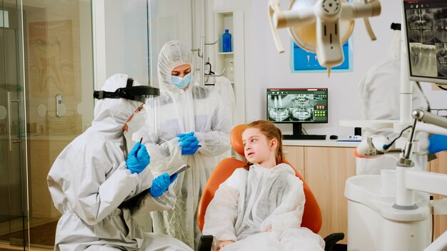 Dentist doctor in coverall talking with girl patient during global coronavirus pandemic writing on clipboard. Nurse and orthodontic doctor wearing face shield, protection suit, mask and gloves.