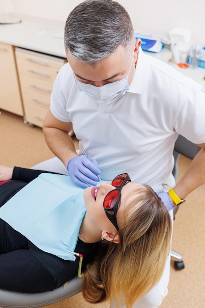 歯科医の医者は患者の歯を見て、口の近くに歯科用器具を持っていますアシスタントは医者を助けます彼らはマスクと手袋で白いユニフォームを着ます歯科医の歯科医院