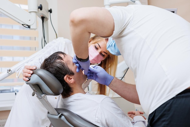 Foto il medico dentista guarda i denti del paziente e tiene gli strumenti dentali vicino alla bocca l'assistente aiuta il medico indossano uniformi bianche con mascherine e guanti dentista studio dentistico