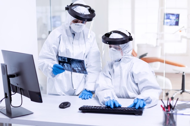 Dentist doctor and nurse in ppe suit against covid-19 and nurse holding teeth x-ray. Medical specialist wearing protective gear against coronavirus during global outbreak looking at radiography in den