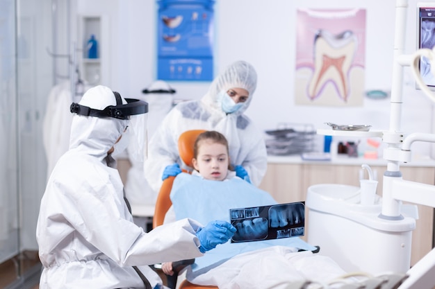 Dentista vestito con tuta in dpi che esamina la radiografia panoramica della bocca del paziente della bambina. stomatologo in tuta protettiva per coroanvirus come precauzione di sicurezza tenendo i raggi x dei denti dei bambini durante la consultazione