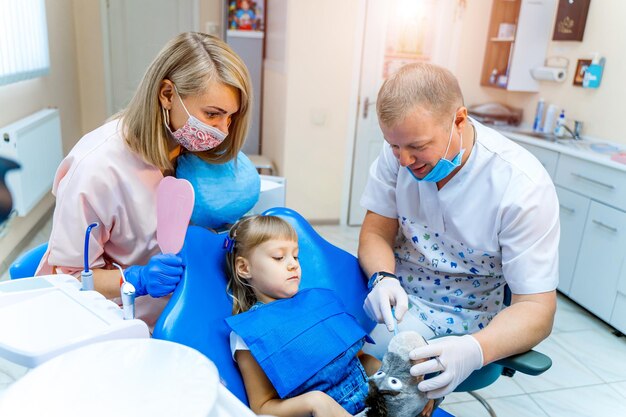 Dentist examining little girl's teeth in the stomatology chair with assistant's help Doctor holds a soft toy Selective focus Pediatric stomatology concept