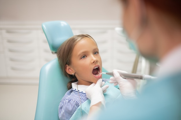 Dentist examining teeth. Beautiful blue-eyed girl opening mouth while dentist examining teeth