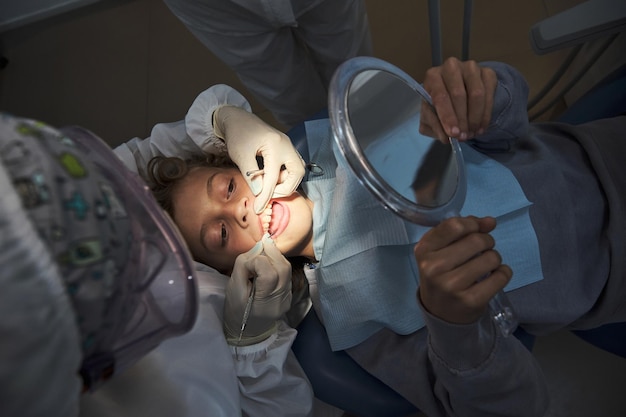 Dentist examining teeth of boy holding mirror in hospital