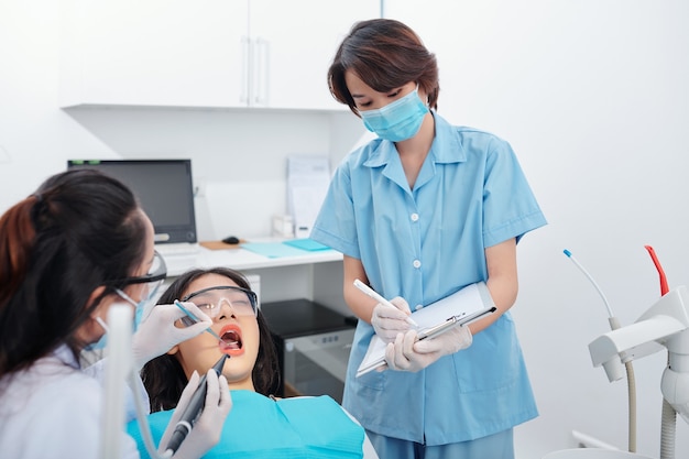 간호사가 문서에 메모를 할 때 여성 환자의 치아를 검사하는 치과 의사