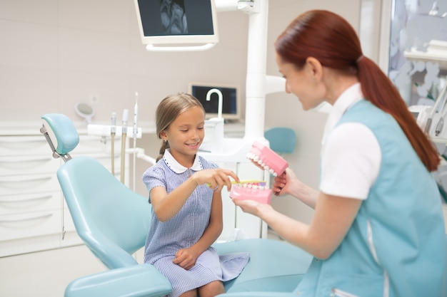 Dentist giving lesson. Red-haired professional child dentist giving girl a lesson of teeth brushing