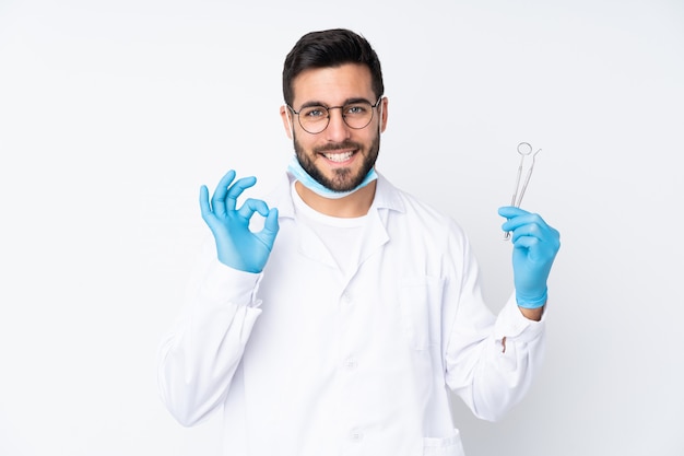 Dentist man holding tools on white wall showing an ok sign with fingers