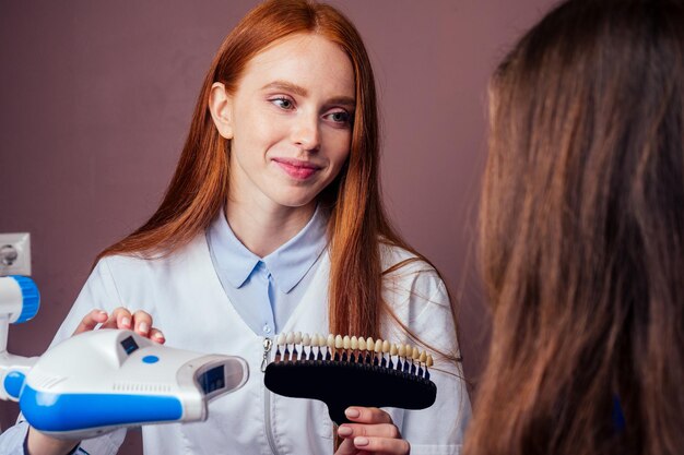 Photo dentist matching teeth color of beautuful red haired ginger woman with palette in clinic, teeth whitening concept.