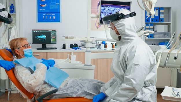 Dentist nurse in coverall putting dental bib to old woman before stomatological examination during covid-19 pandemic. Assistant and orthodontic doctor wearing protection suit, face shield mask gloves