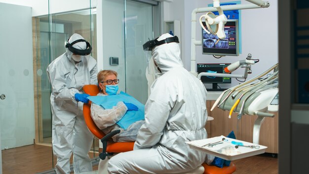 Dentist nurse in coverall putting dental bib to old woman before stomatological examination during covid-19 pandemic. Concept of new normal dentist visit in coronavirus outbreak wearing protective sui