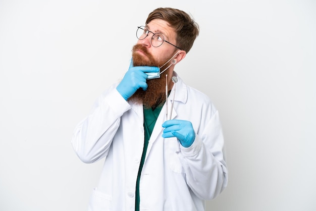 Dentist reddish man holding tools isolated on white background having doubts