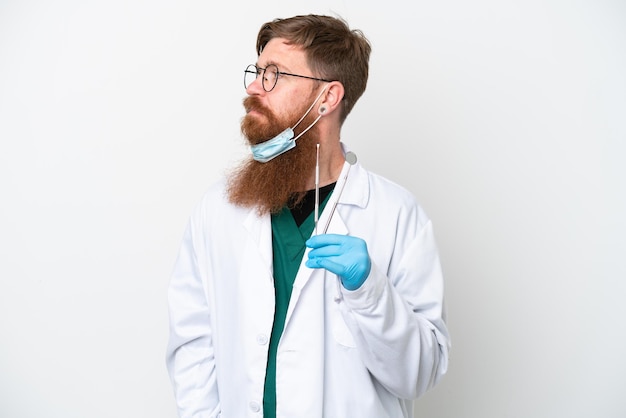 Dentist reddish man holding tools isolated on white background looking to the side