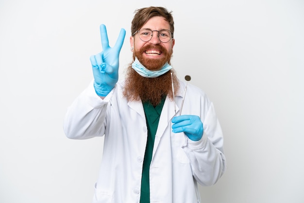 Dentist reddish man holding tools isolated on white background smiling and showing victory sign