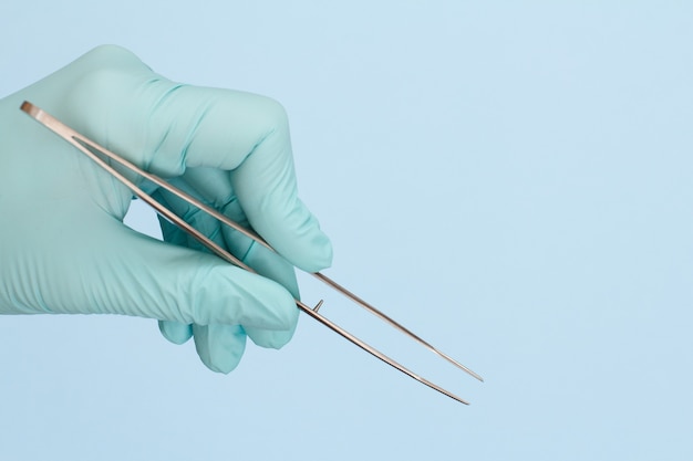 Dentist's hand in latex glove with tweezers on blue background. Medical tools concept.