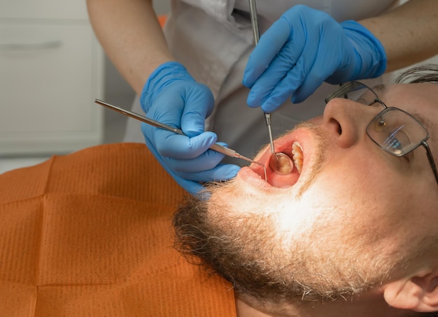 Le mani del dentista tengono gli strumenti per esaminare i denti di un uomo bianco con gli occhiali e la barba