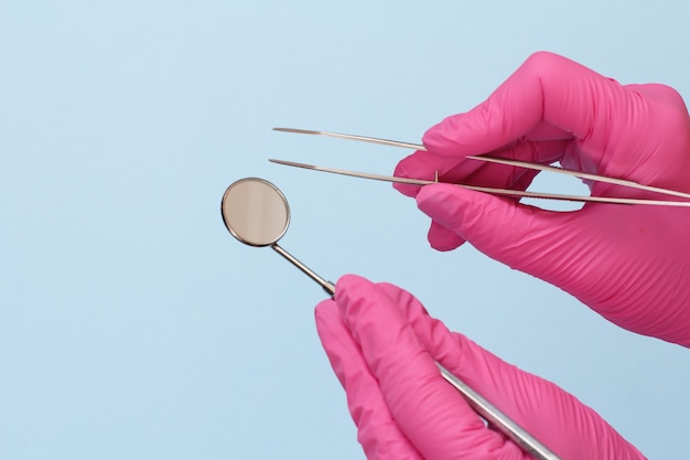 Dentist's hands in pink latex gloves with tweezers and mouth mirror on blue background. Medical tools concept.