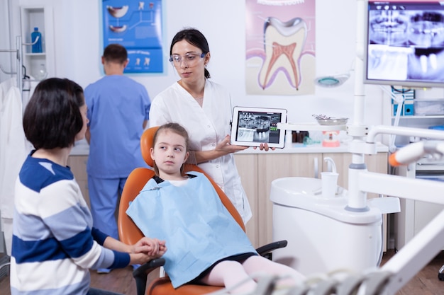 Dentist specialist discussing about jaw digital radiography of little girl patient. Stomatologist explaining teeth diagnosis to mother of child in health clinic holding x-ray.