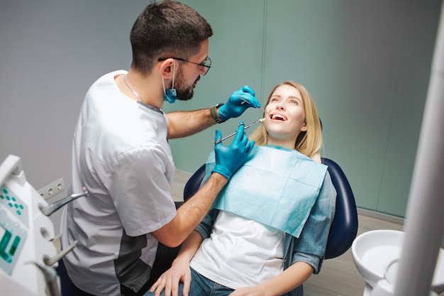 Dentist stand uppon client and using syringe for putting pain killer. Client sits calm in chair and keep mouth opened. Careful dentist. Patient client.