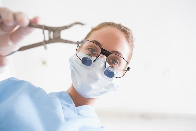 Dentist in surgical mask and dental loupes holding pliers over patient