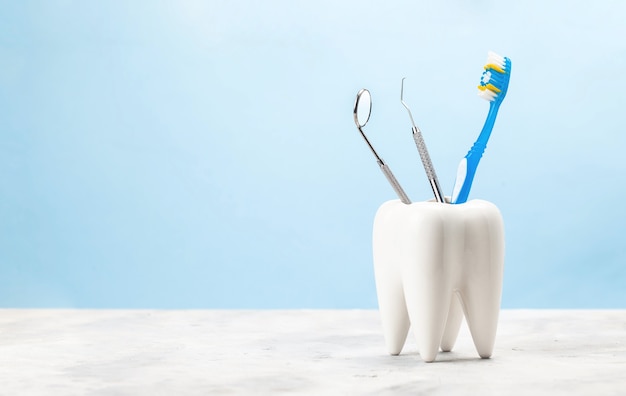 Dentist toothbrush and steel tools mirror and hook in tooth shaped glass. Blue background.