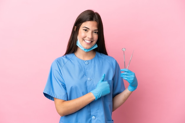 Dentist woman holding tools isolated on pink background giving a thumbs up gesture