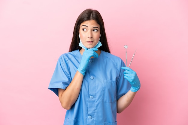 Женщина стоматолога, держащая инструменты, изолированные на розовом фоне, сомневаясь и думая