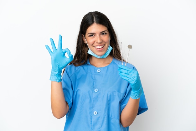 Dentist woman holding tools over isolated white background showing ok sign with fingers