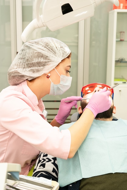 Dentist young woman treats a patient a man. The doctor uses disposable gloves, a mask and a hat. The dentist works in the patient's mouth, uses a professional tool