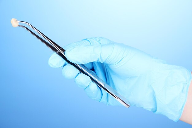 Dentists hands in medical gloves holding tweezers on blue background