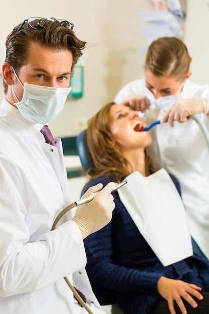 Dentistshis surgery holds a drill and looking at the viewer, in the  his assistant is giving a female patient a treatment