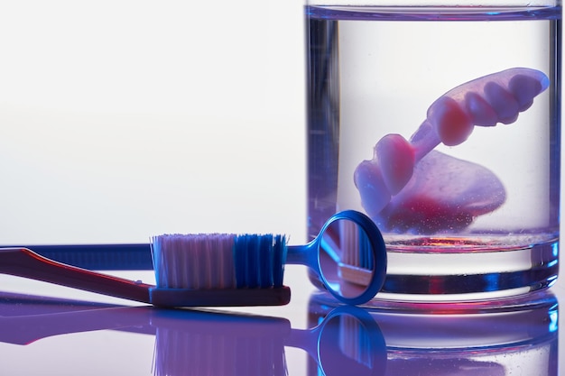 Denture in a glass of water with toothbrush and mirror dental\
prosthesis care