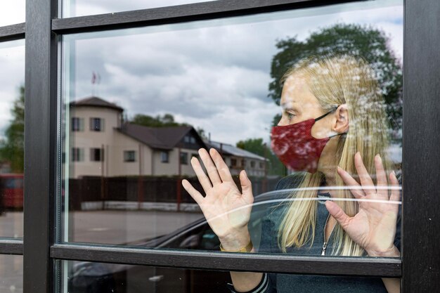 Foto donna depressa con la maschera che guarda fuori dalla finestra che rimane a casa in quarantena a causa del lockdown covid-19