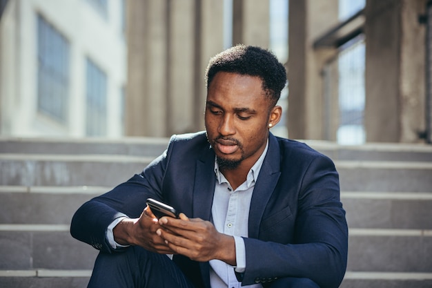 Подавленный афро-американский бизнесмен, читающий плохие новости по мобильному телефону, разочарованный и грустный, сидя на лестнице в деловом костюме