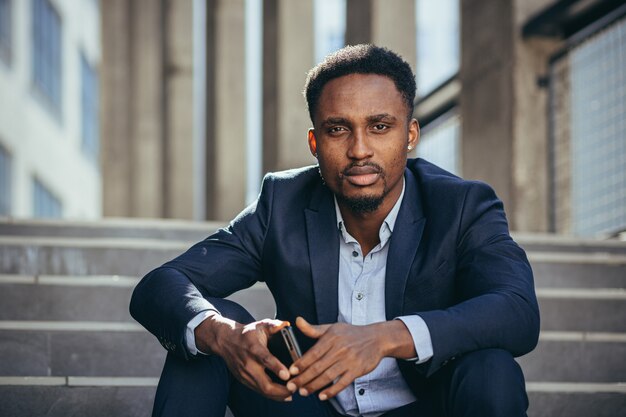 Depressed african american businessman reading bad news from cellphone, frustrated and sad sitting on stairs in business suit