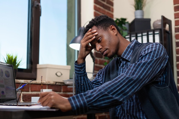 Depressed african american office worker feeling tired because of startup low profit analytics on laptop display. Marketing specialist having a headache sitting at desk working with business charts.