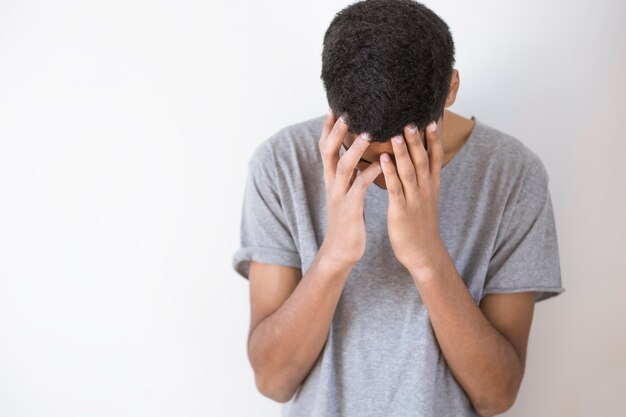 Giovane afroamericano depresso su fondo bianco. un triste ragazzo nero si copre la faccia con le mani.