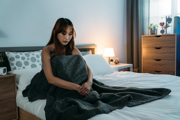 不眠症の落ち込んだアジアの女の子は、夜明けの寝室で孤独を感じながら膝に手を置いてベッドに座っています。
