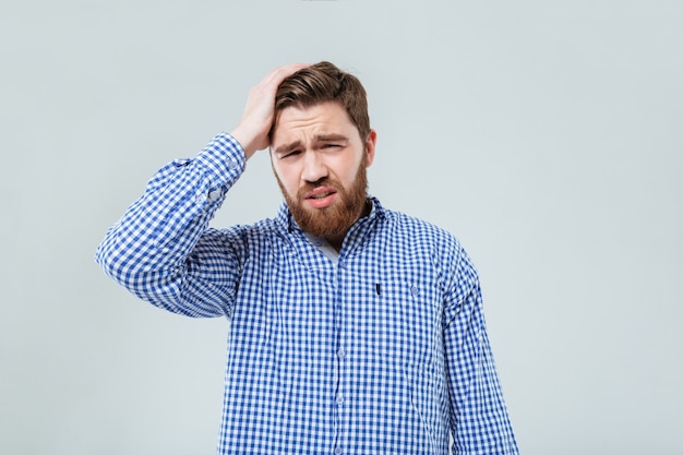 Depressed bearded young man standing and having headache over white wall