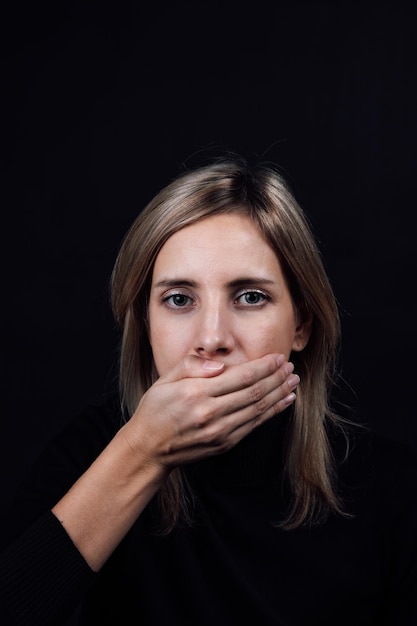 Depressed blonde woman in black blouse with hand covering mouth and making her be silent on black background Victim of physical and psychological abuse Gaslighting Relative aggression