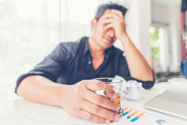 Foto uomo d'affari depresso con un bicchiere di whisky mentre è seduto con la testa tra le mani alla scrivania dell'ufficio