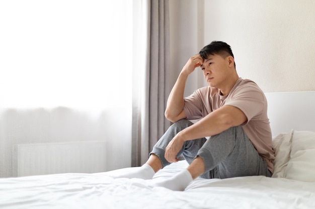 Depressed chinese middle aged man sitting on bed copy space