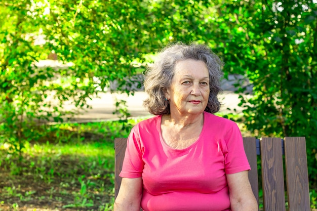 Depressed elderly woman thoughtful while sitting on a park bench on a summer sunny day. A mature woman in a bright T-shirt thinks about sad things. Psychology. Serious and thoughtful expression