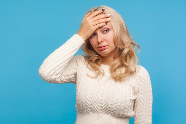 Photo depressed hopeless woman with curly blond hair making facepalm gesture, shame, getting into trouble, making mistake. indoor studio shot isolated on blue background