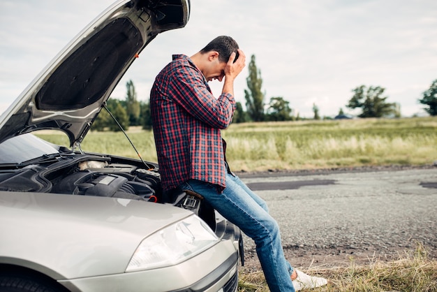 Depressed man sitting on a hood of broken car. Vehicle with open hood on roadside