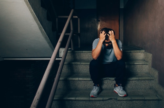 Photo depressed man sitting on the stairs in building and holding his forehead while having headache.