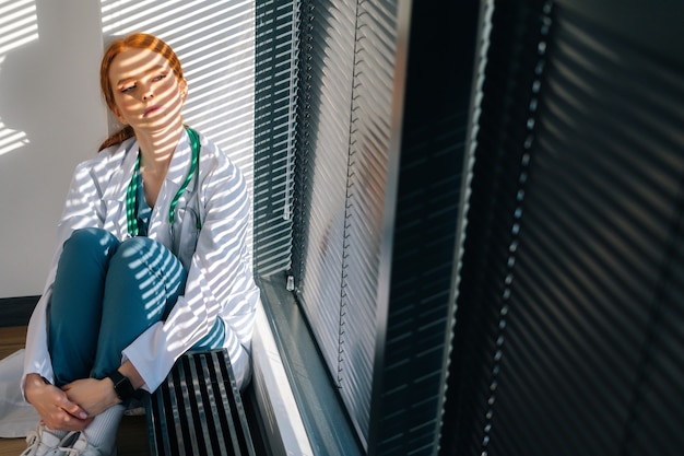 Depressed sad young female doctor in white coat sitting on floor hugging legs with hands by window